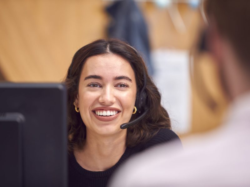 Businesswoman In Cubicle Wearing Headset Talking To Colleague In Busy Customer Services Centre