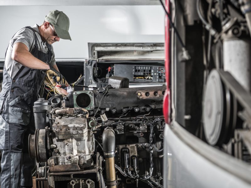 Professional Caucasian Automotive Mechanic in His 40s Repairing Powerful Heavy Duty Truck or Bus Diesel Engine Inside Truck Service Center. Transportation Industry Theme.